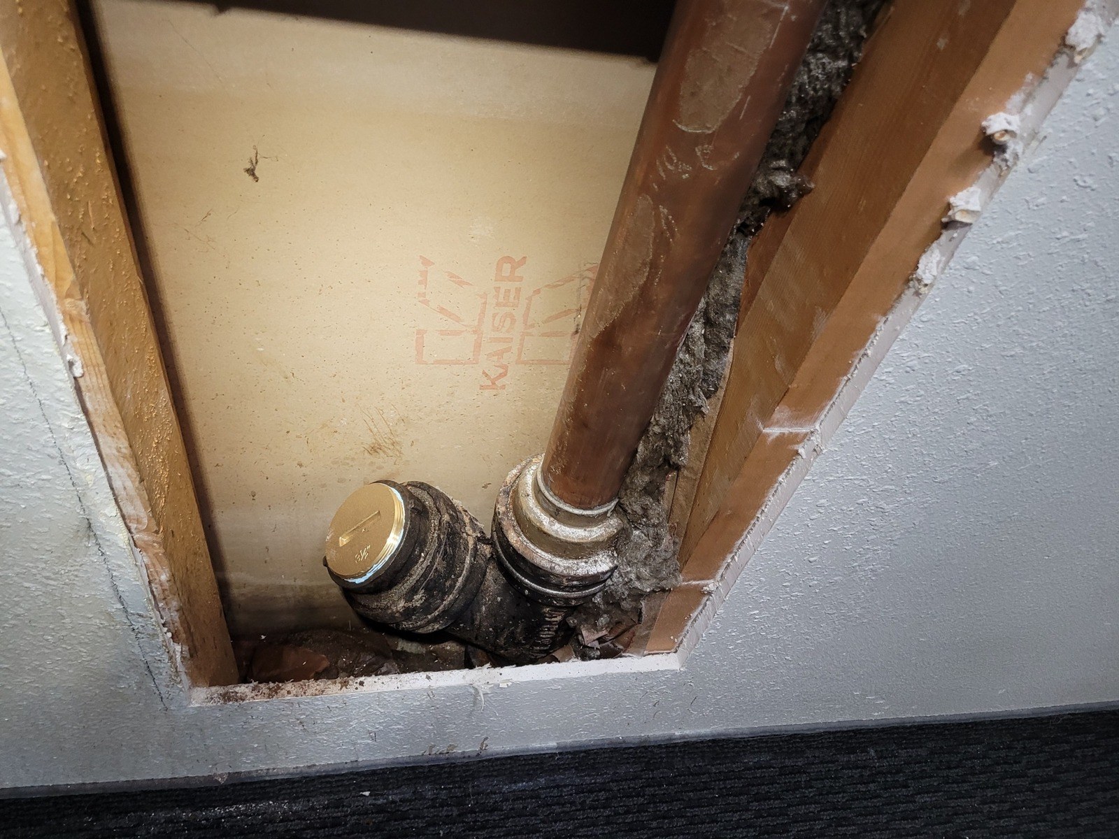 Opened wall showing a cast-iron drain fitting with a new brass cleanout plug beside a vertical copper pipe, with the drywall backing stamped by its manufacturer