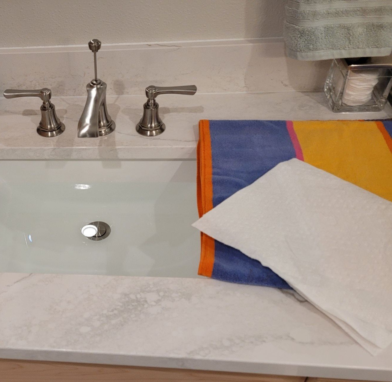 A brushed-nickel widespread bathroom faucet with lever handles on a white marble-look vanity top, with the undermount sink filled with water and a sheet of tissue paper resting on a towel beside it