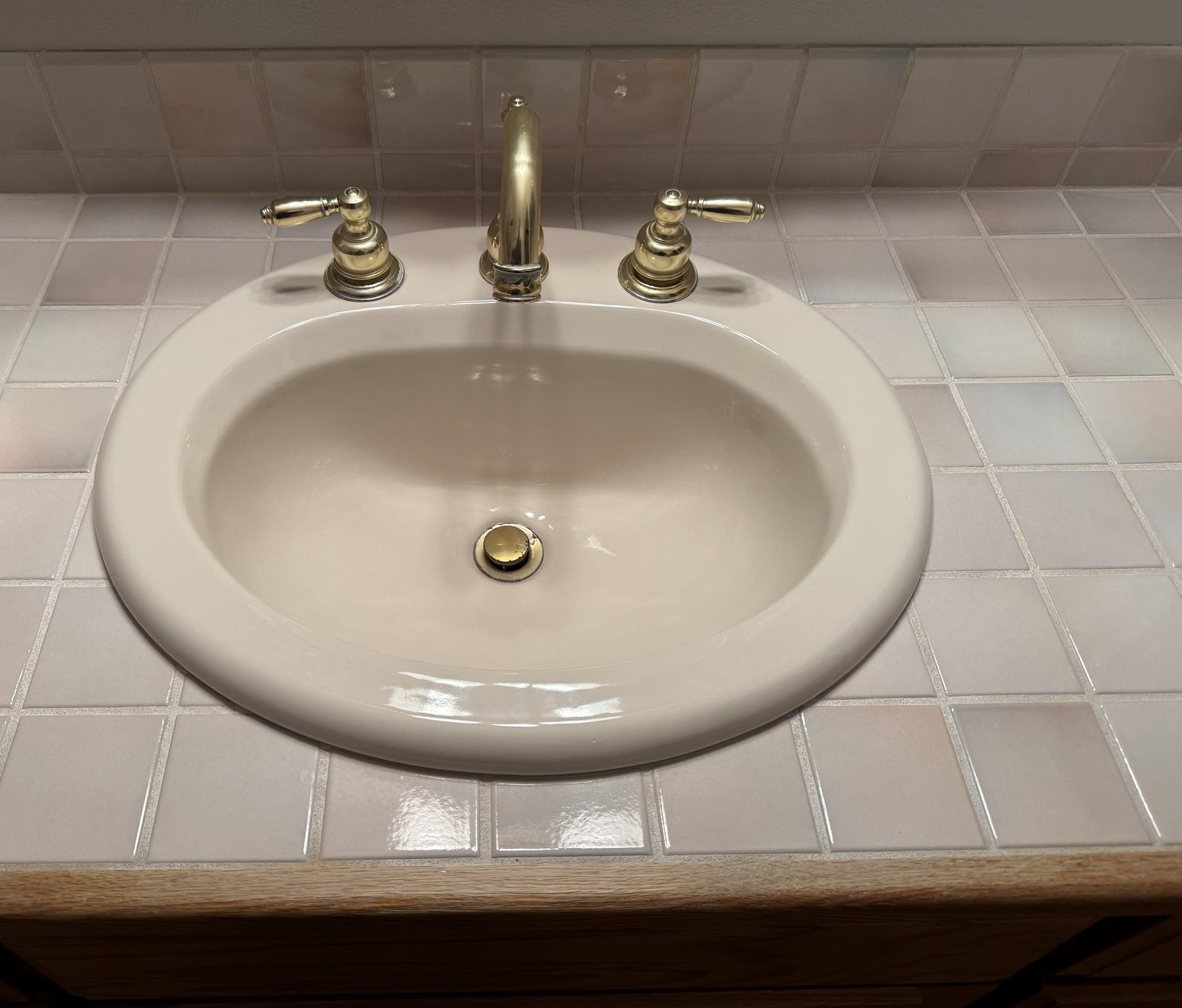 Old polished-brass widespread faucet with worn lever handles and corrosion at the base of the spout, on an almond oval sink set in pale pink tile