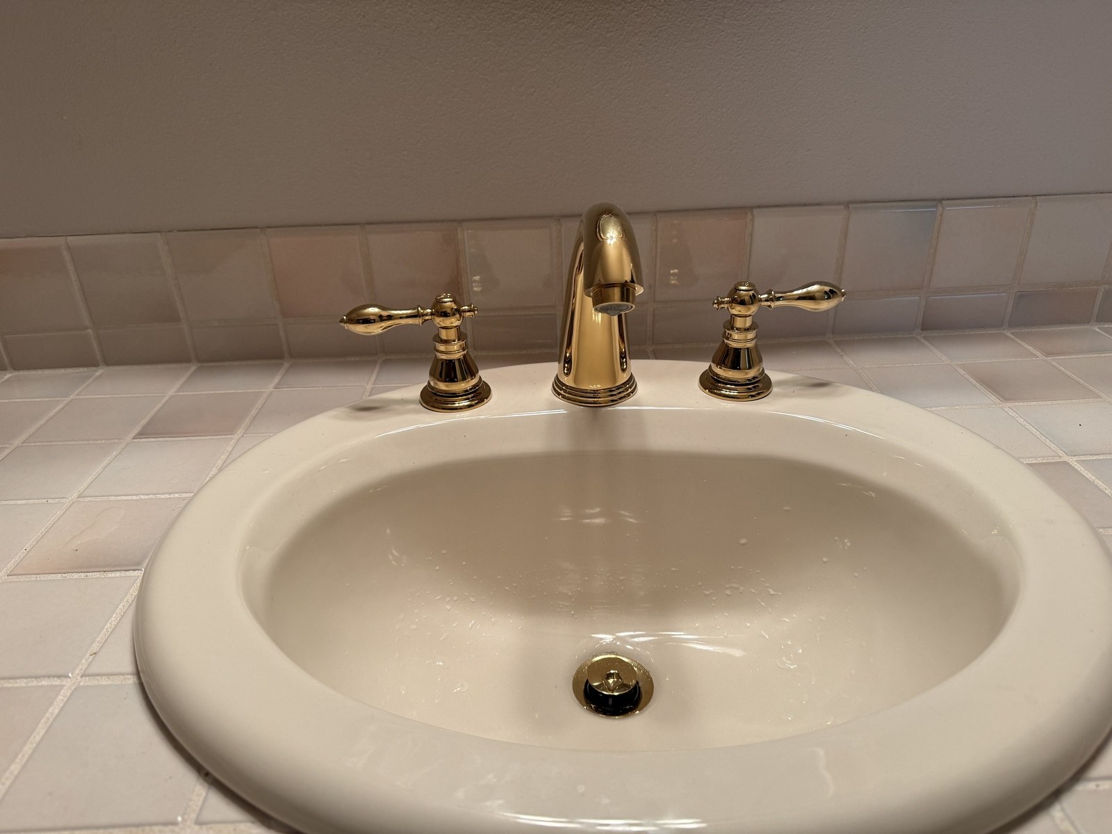 Polished-brass widespread bathroom faucet with lever handles on an almond oval sink set in pale pink tile, the basin still wet from testing