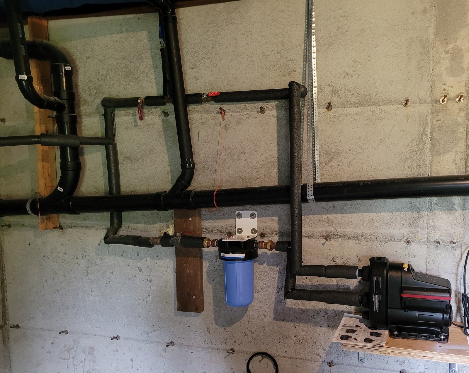 Black Grundfos booster pump on a plywood shelf against a concrete foundation wall, its new supply lines sleeved in black foam insulation beside a blue whole-house filter