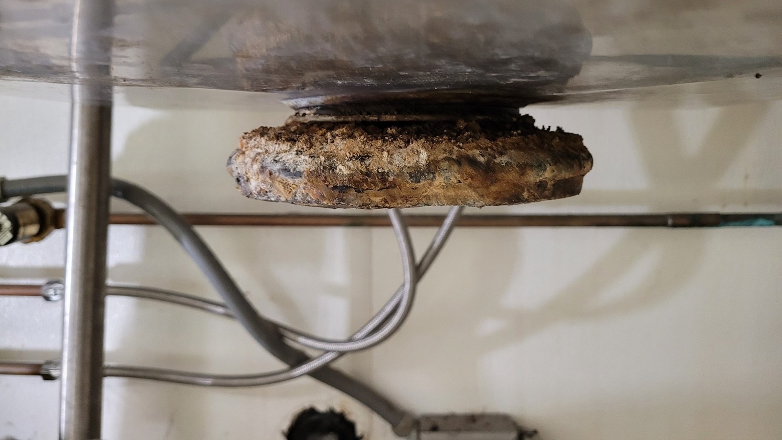 Underside of a stainless sink bowl with a corroded, crusted disposer collar hanging from the drain opening above braided supply lines