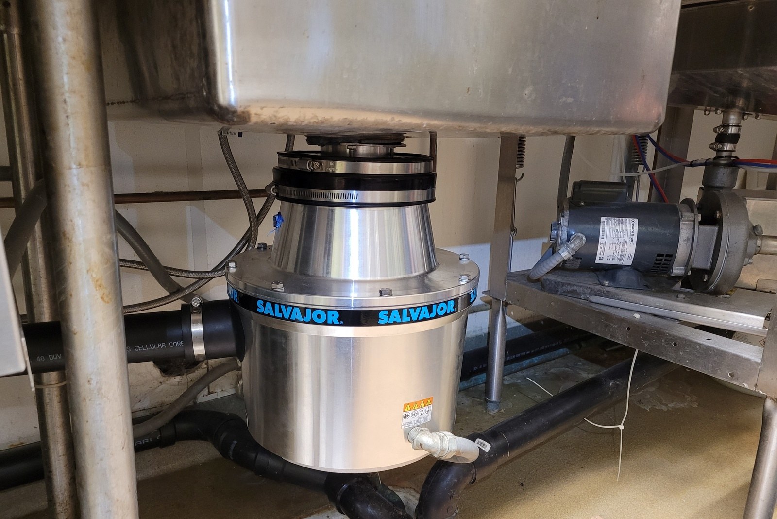 New stainless Salvajor commercial food disposer with its blue-lettered band, mounted under a stainless dish sink with its black drain line running down to a floor sink