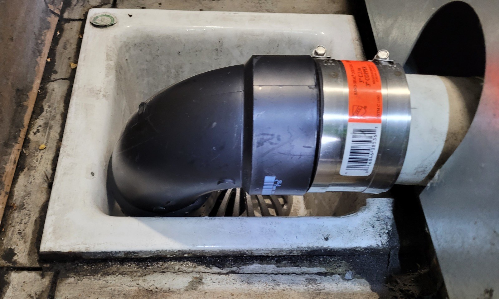 A black plastic elbow joined by a banded coupling to the machine's discharge pipe, turning down into a white square floor sink set in a concrete floor beside the steel side of the extractor