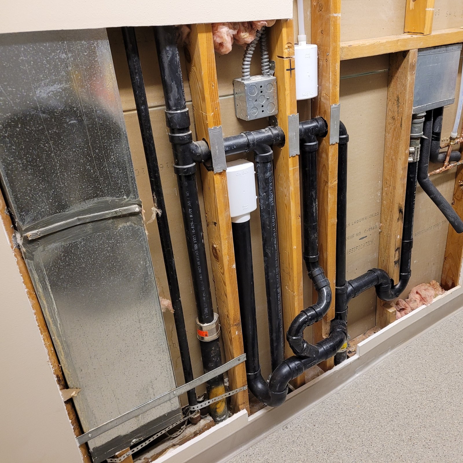 Opened laundry-room wall showing new black ABS drain and vent lines, two recessed white washer boxes with supply valves, copper stubs and an electrical box between wood studs