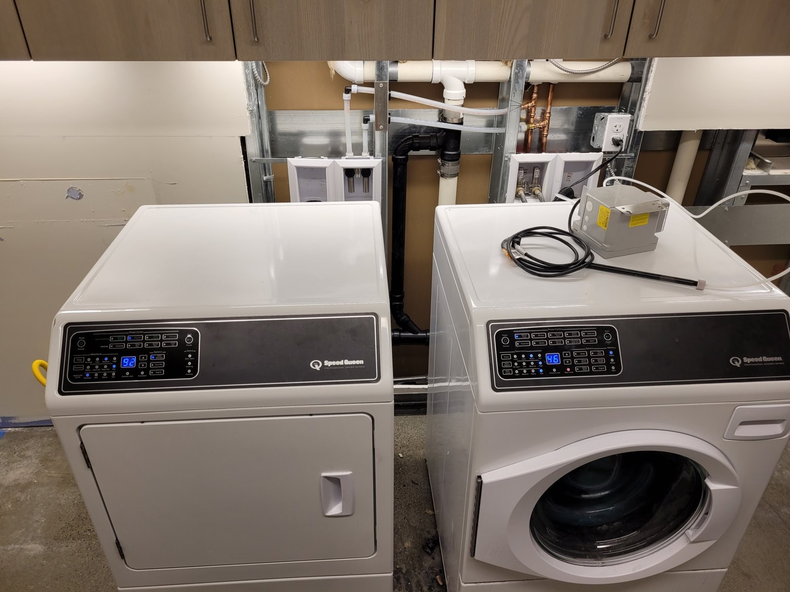 Two new white commercial front-load laundry machines set against an opened wall where new recessed washer boxes, black ABS drain lines and copper supply stubs have been built between the studs
