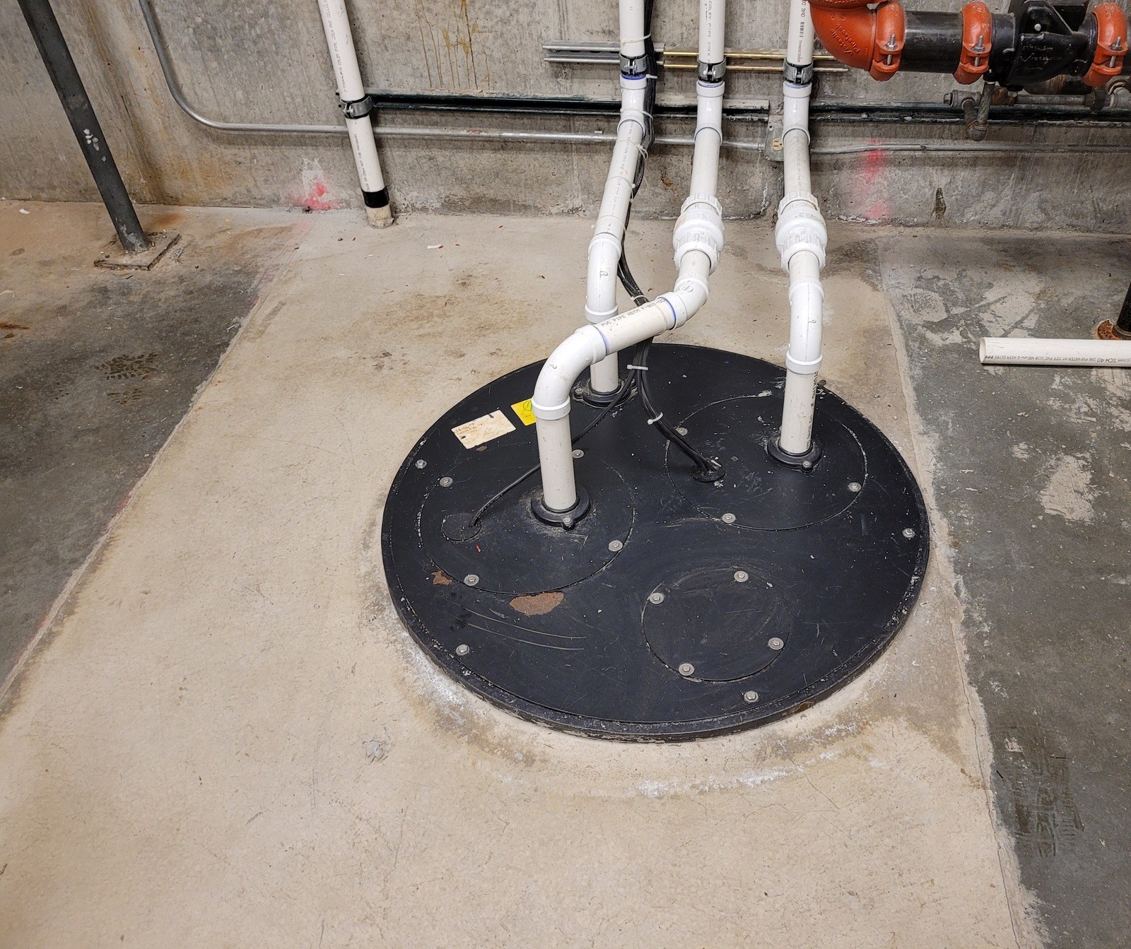 The same sewage chamber lid in a concrete floor, with three new white PVC discharge pipes rising from it, unions near the floor and lengths of spare pipe lying beside it