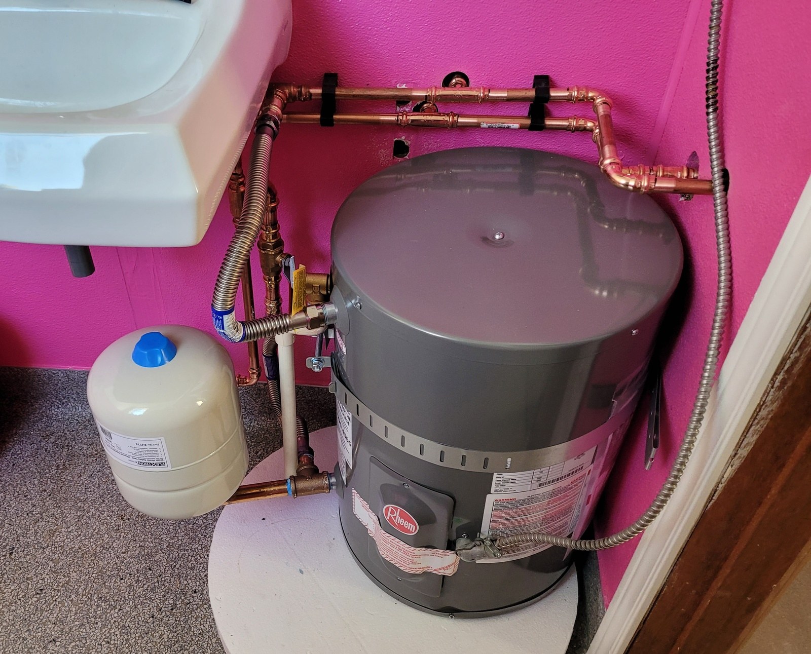 Small gray Rheem electric water heater on a white foam pad beside a wall-hung sink, with new copper press-fitted piping, an expansion tank and ball valves against a pink wall