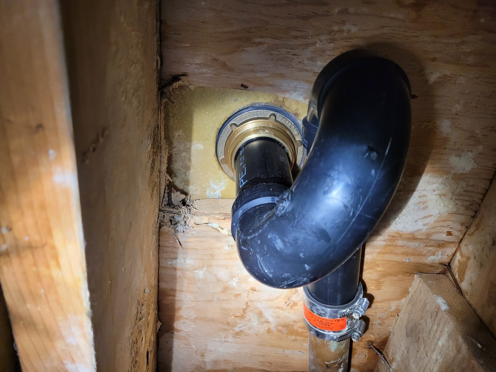New brass shower drain body and black ABS trap seen from below through an opening in a ceiling, between wood framing