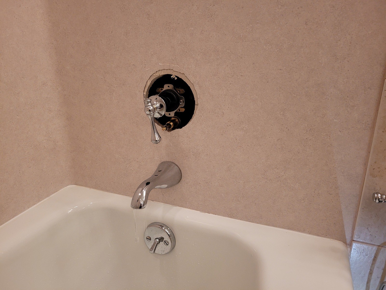 Kohler tub and shower valve with its trim plate removed, showing the new cartridge and lever handle in a round wall opening in a beige cultured-marble surround, with the chrome tub spout running into a white tub below