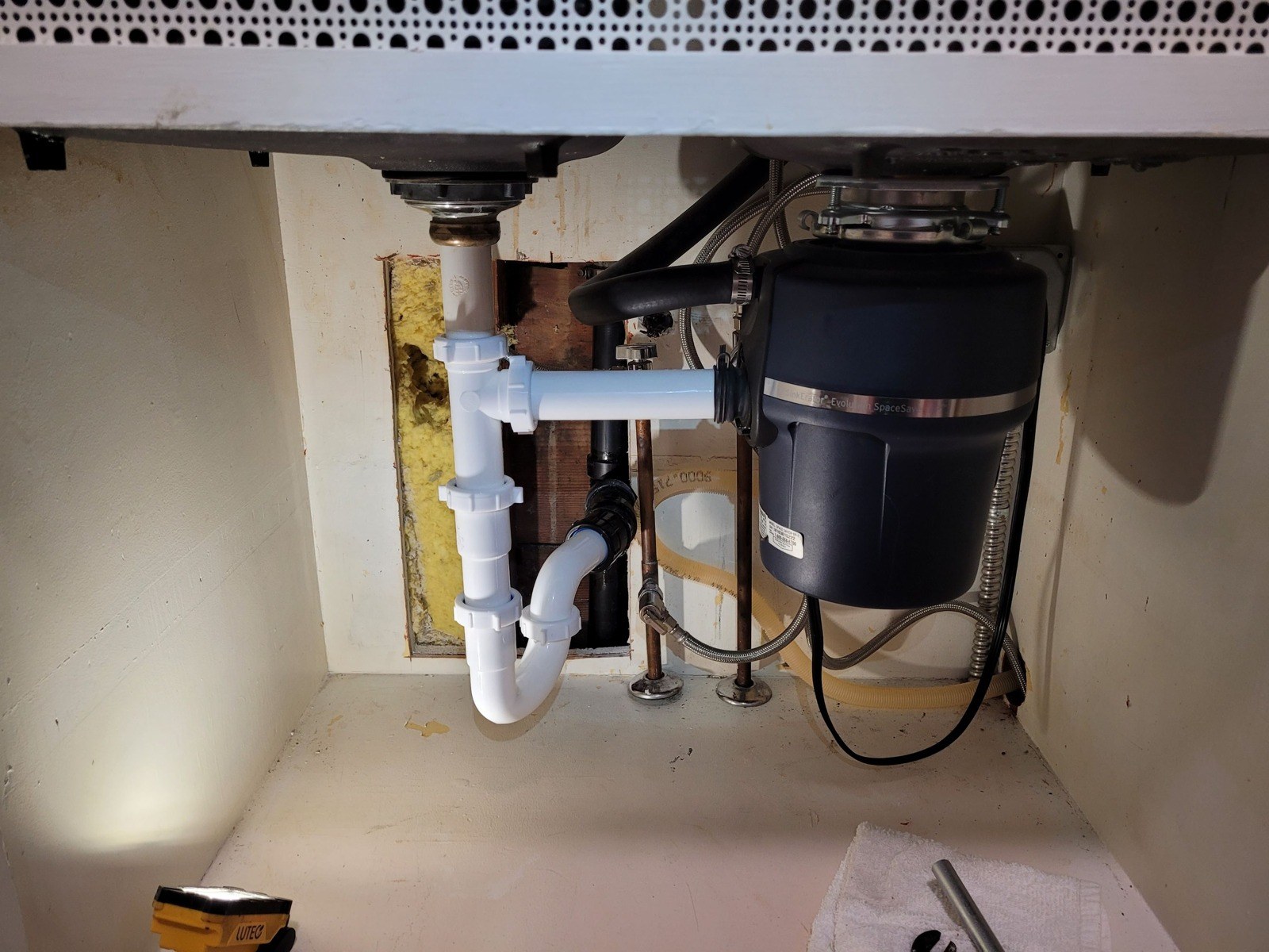 Under a kitchen sink, a new white P-trap connected to a garbage disposal, with the cabinet back opened to show yellow insulation and new black ABS drain pipe
