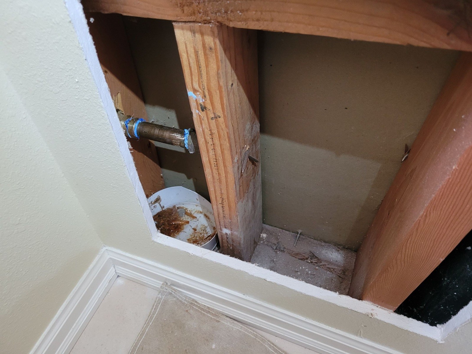 Rectangular opening cut in a painted wall at baseboard height, exposing a wood stud and a threaded brass pipe stub wrapped in blue tape, with a bucket below