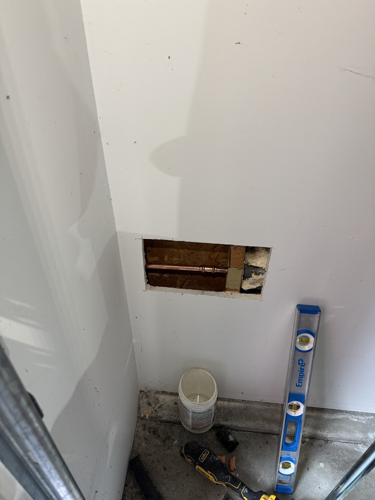 Rectangular access opening cut in a garage wall exposing the copper supply stub for an outdoor faucet