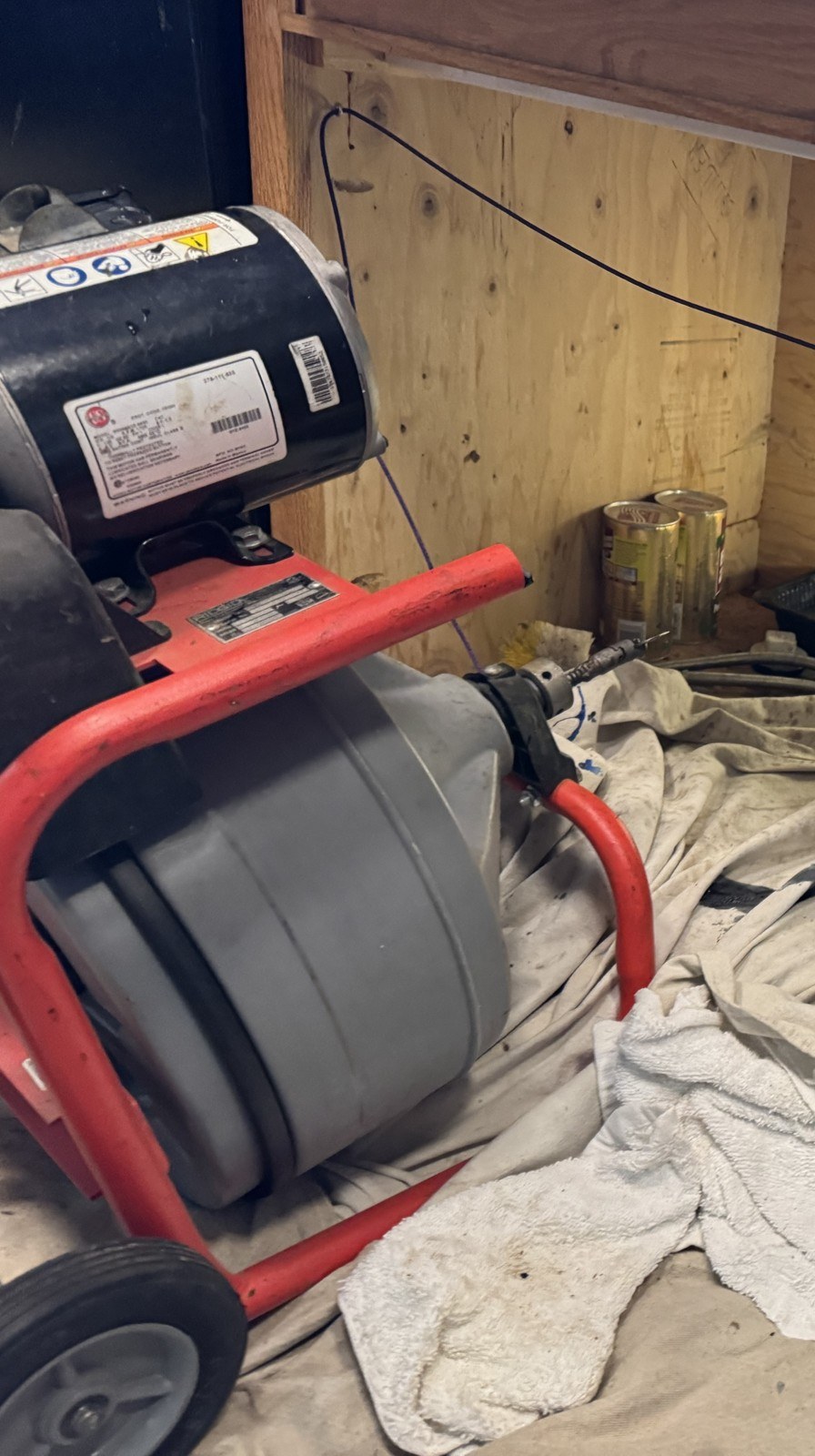 Red drain cleaning machine on its wheeled frame beside an open kitchen sink cabinet, with drop cloths spread on the floor
