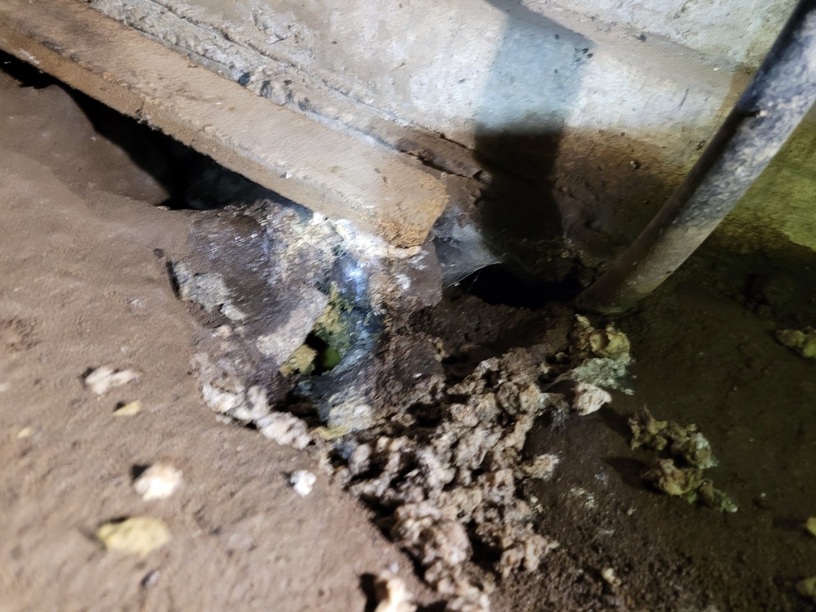 Corroded drain pipe passing into a concrete foundation in a dirty crawlspace, with a failed joint visible