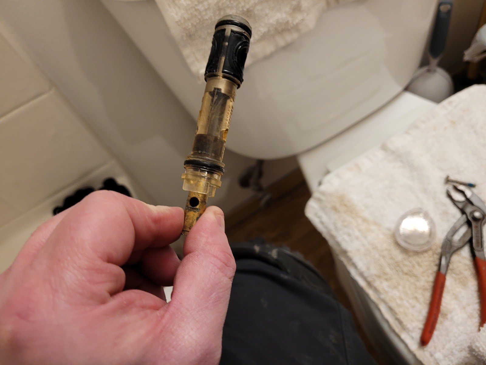 Plumber's hand holding a removed Moen shower cartridge with a brass base, a clear amber body and a black cap, stained brown, over a bathroom floor