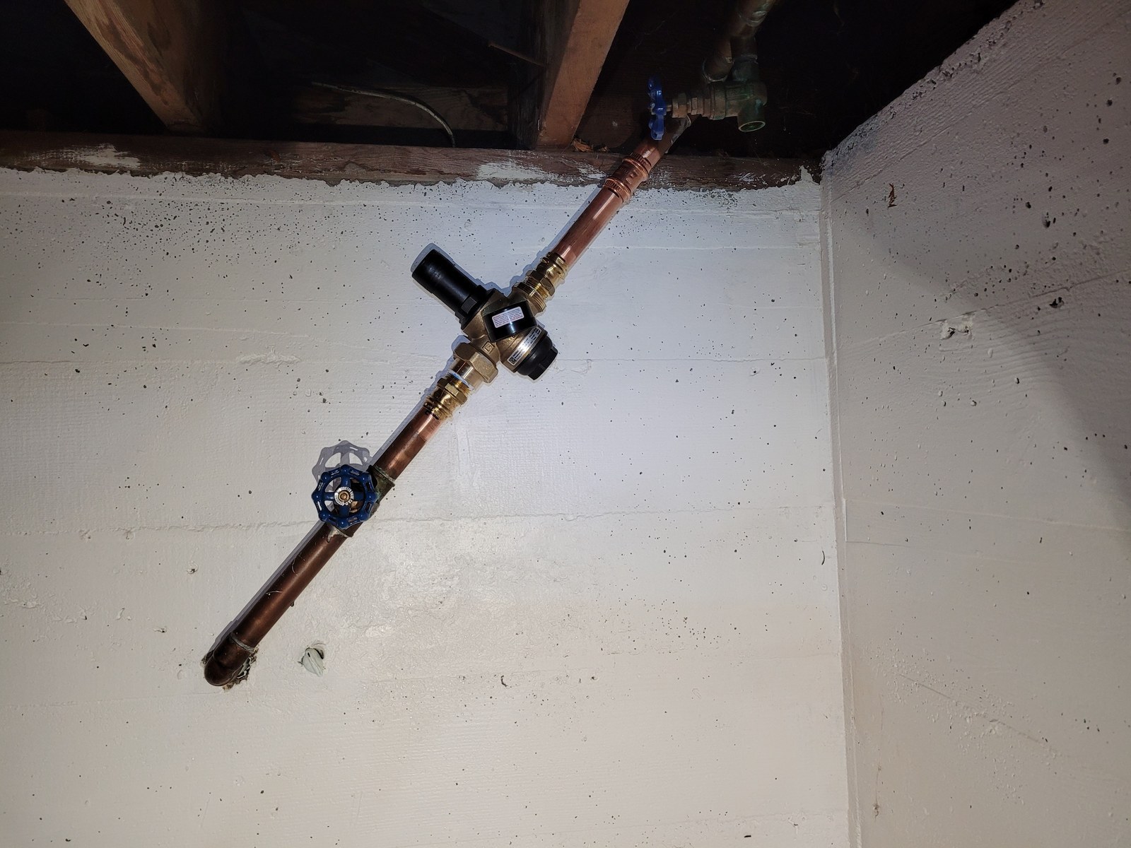 Pressure-reducing valve with a black bell and a blue-handled valve on a copper water main running diagonally down a white-painted basement wall