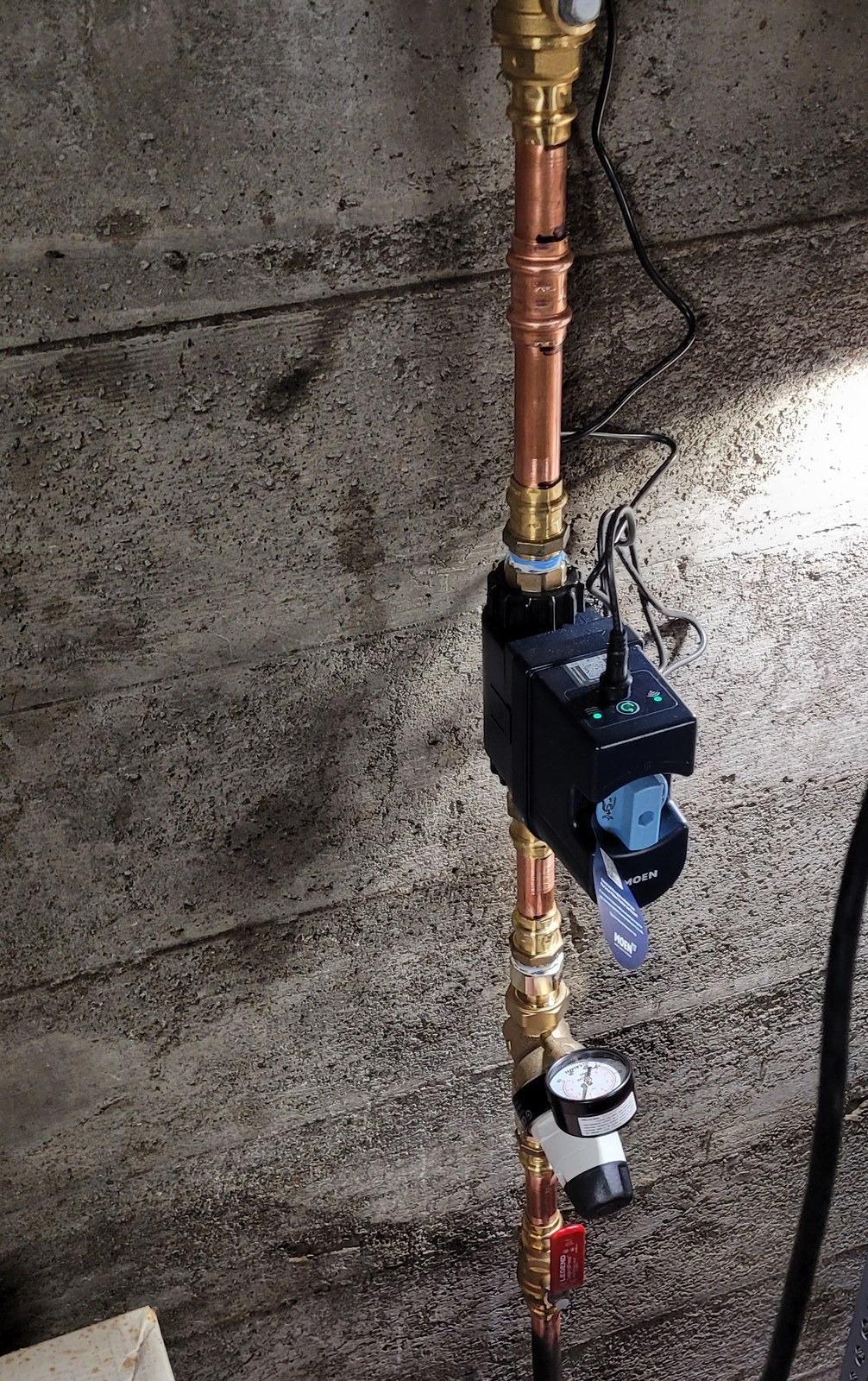 Vertical copper water main on a board-formed concrete wall, with a red-handled ball valve at the bottom, a pressure-reducing valve carrying a small gauge, and a black smart water shutoff above it