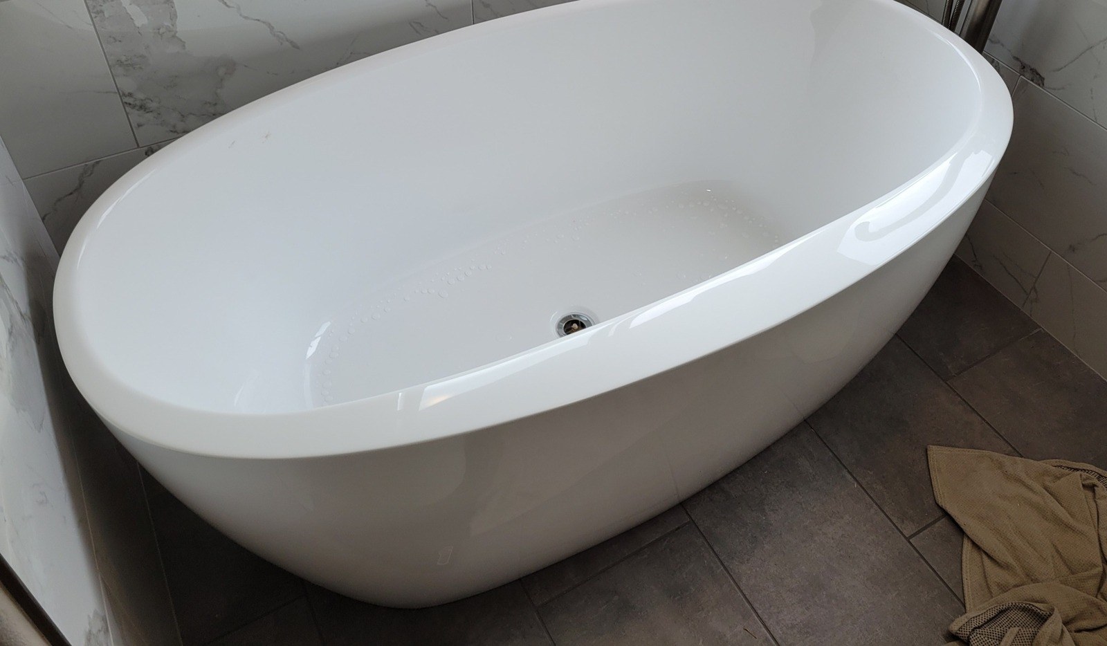 White freestanding oval soaking tub with a brushed-nickel floor-mounted filler and hand shower, set against marble-look wall tile on a dark plank-tile floor