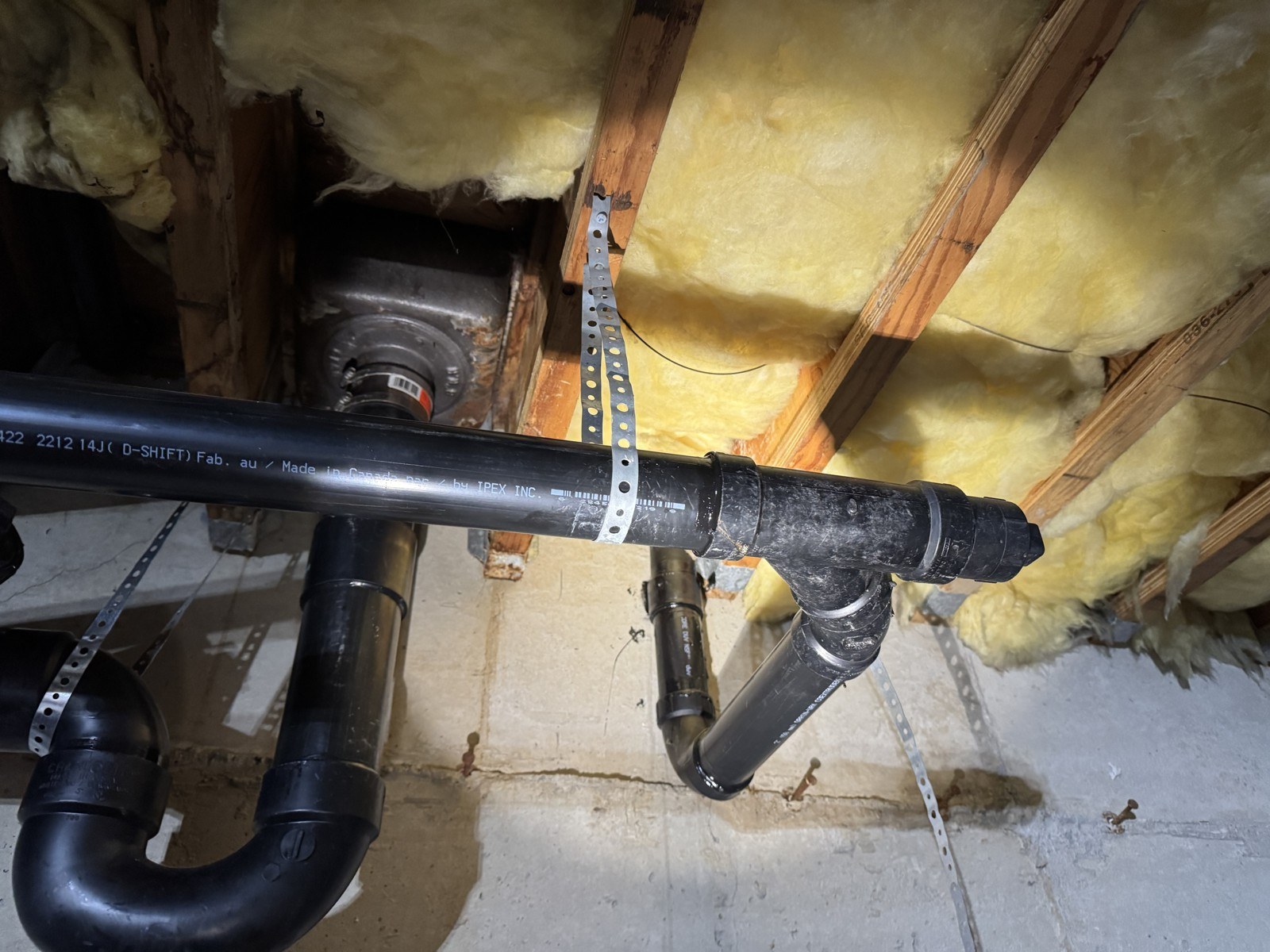 Black ABS drain piping with a P-trap and a sanitary tee hung from metal strapping under a restaurant kitchen floor, with insulation between the joists above