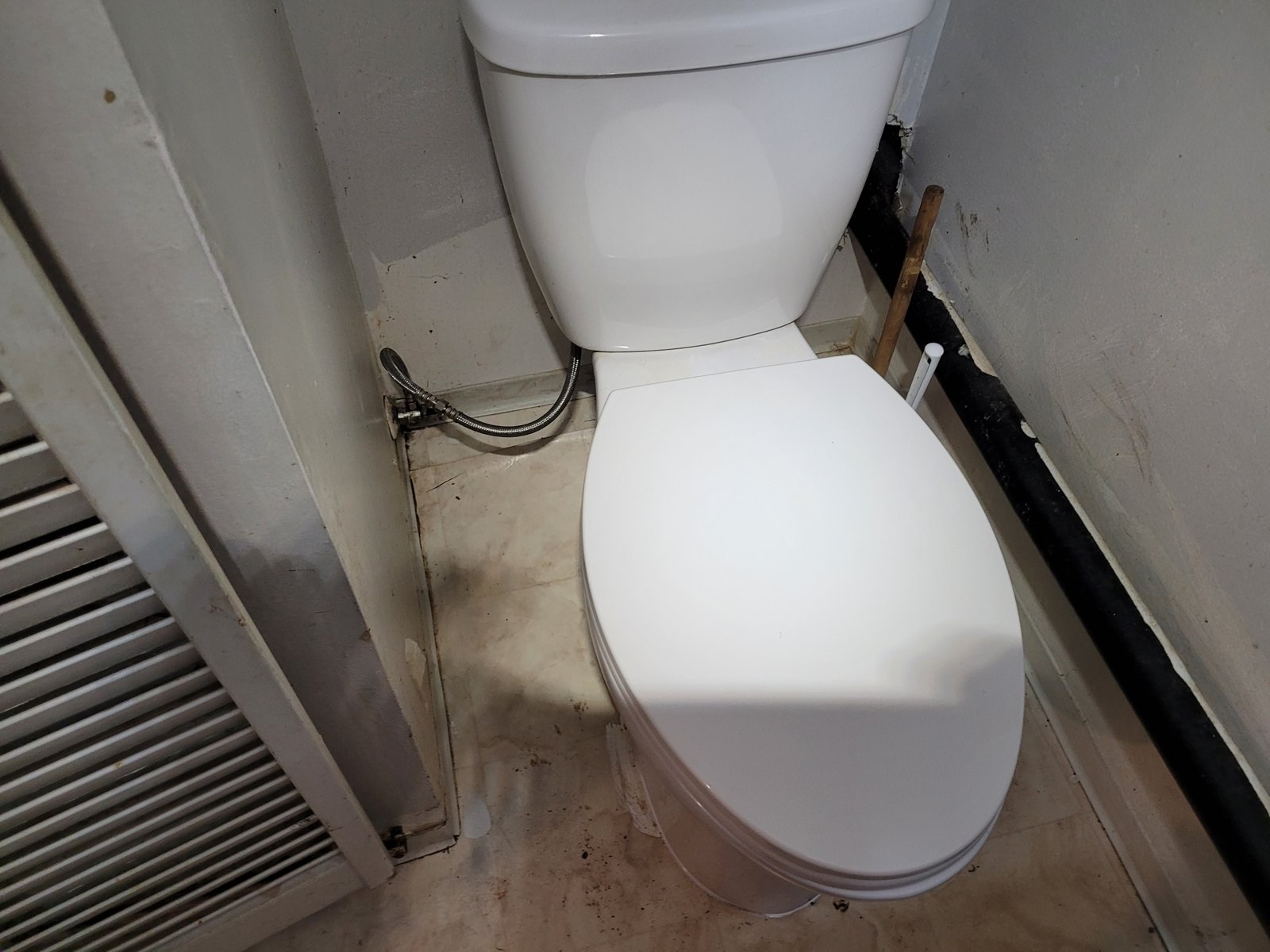 White two-piece toilet with a dual-flush button in a small powder room with a louvered door, a braided supply line and a stained floor around the base