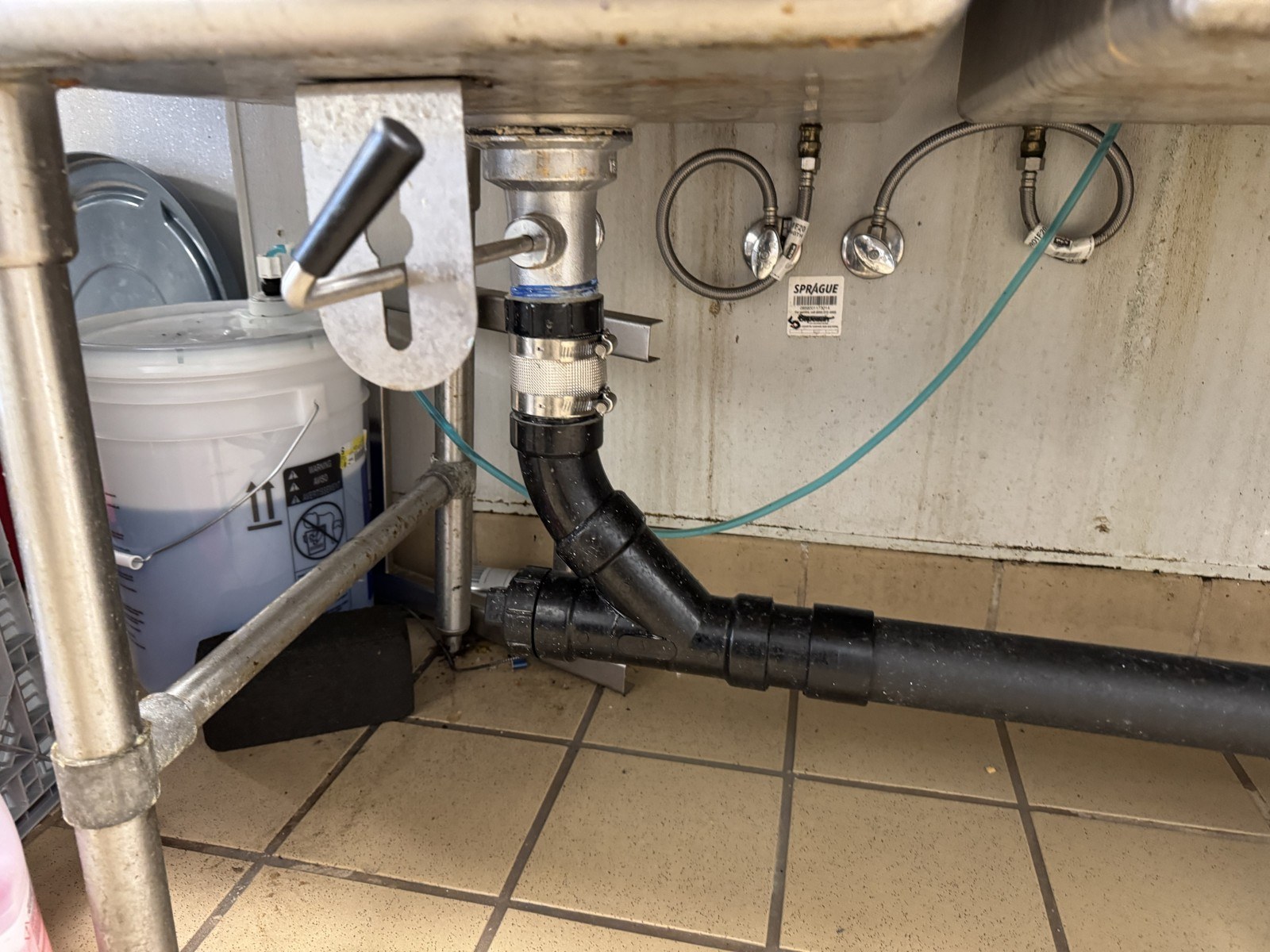 Stainless three-compartment sink in a commercial kitchen with its lever waste valve and black ABS drain running to a wye on the tiled floor