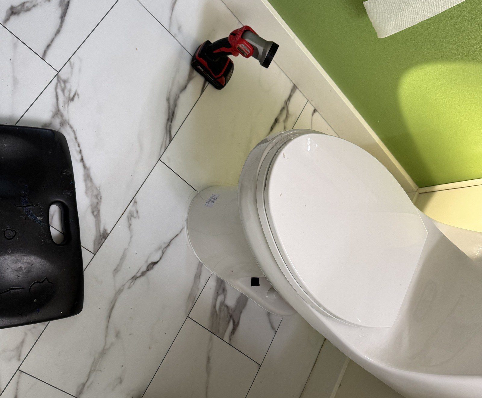 Looking down at the base of a white one-piece toilet where it meets marble-look floor tile, with a red work light and a black kneeling pad on the floor beside it