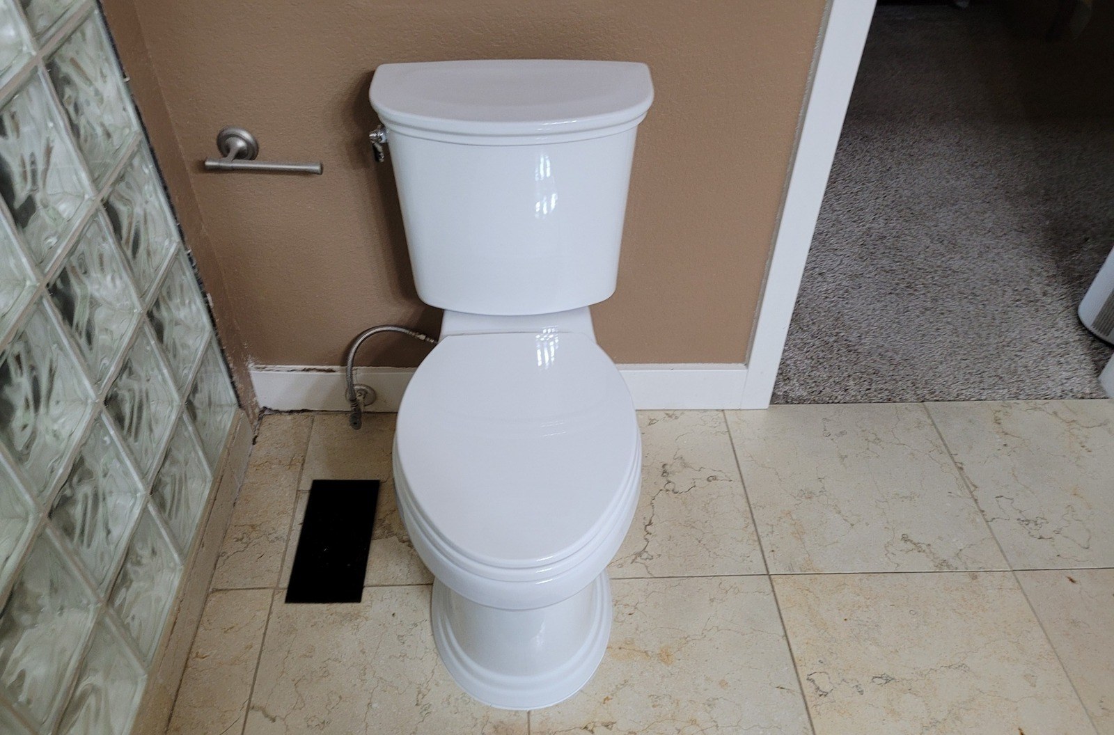 New white skirted two-piece toilet installed beside a glass-block shower wall on beige marble-pattern tile, with a braided steel supply line running to a chrome stop at the baseboard