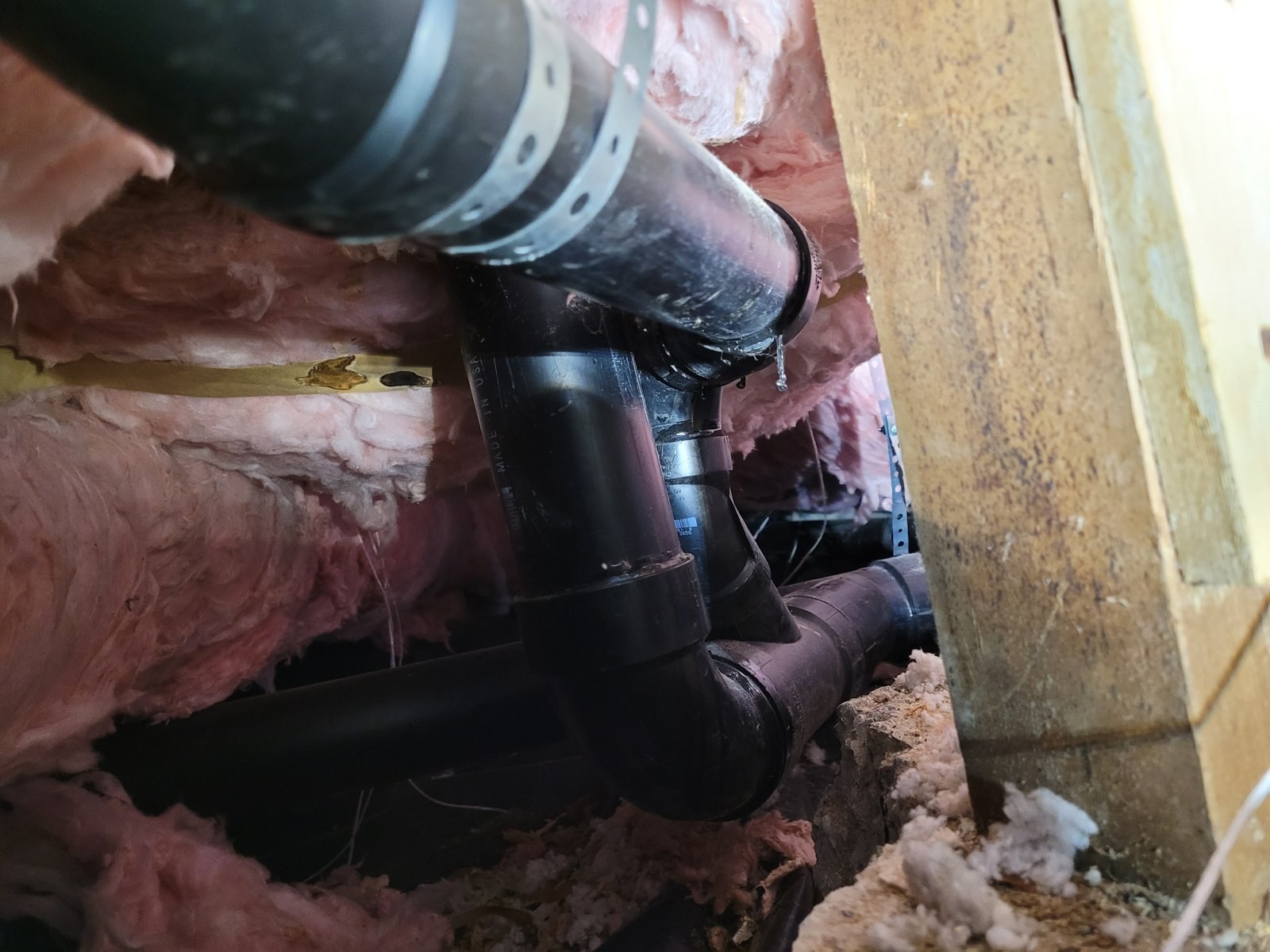 Black ABS drain and vent fittings joined under a house, with pink insulation batts between the floor joists above them
