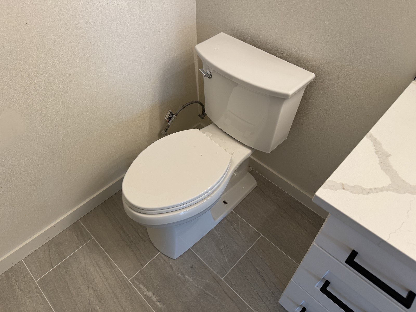 White elongated toilet with a closed lid in a newly tiled bathroom with grey plank tile and a white vanity with a marble-look top