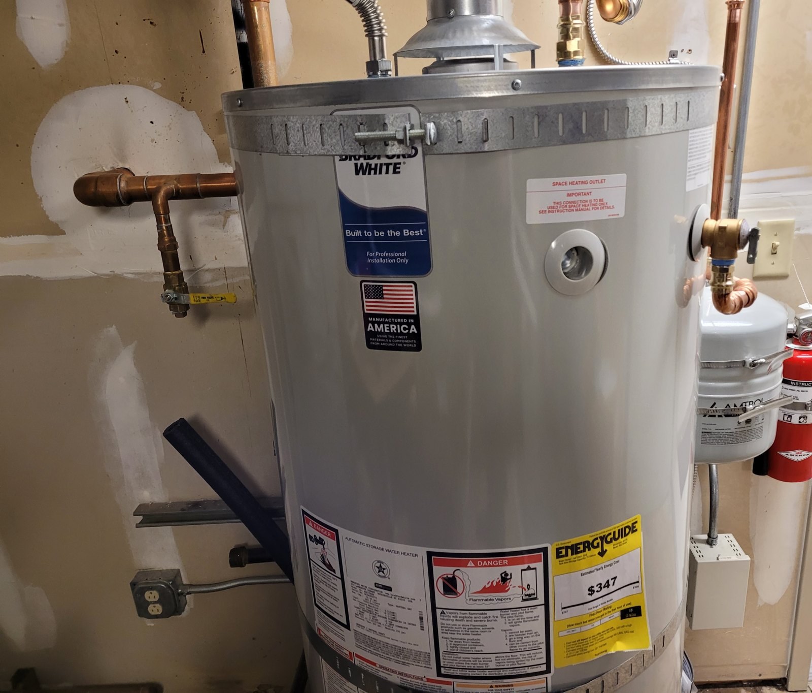 New grey 75-gallon gas water heater with copper piping and a white expansion tank mounted beside it in a mechanical room with unfinished drywall