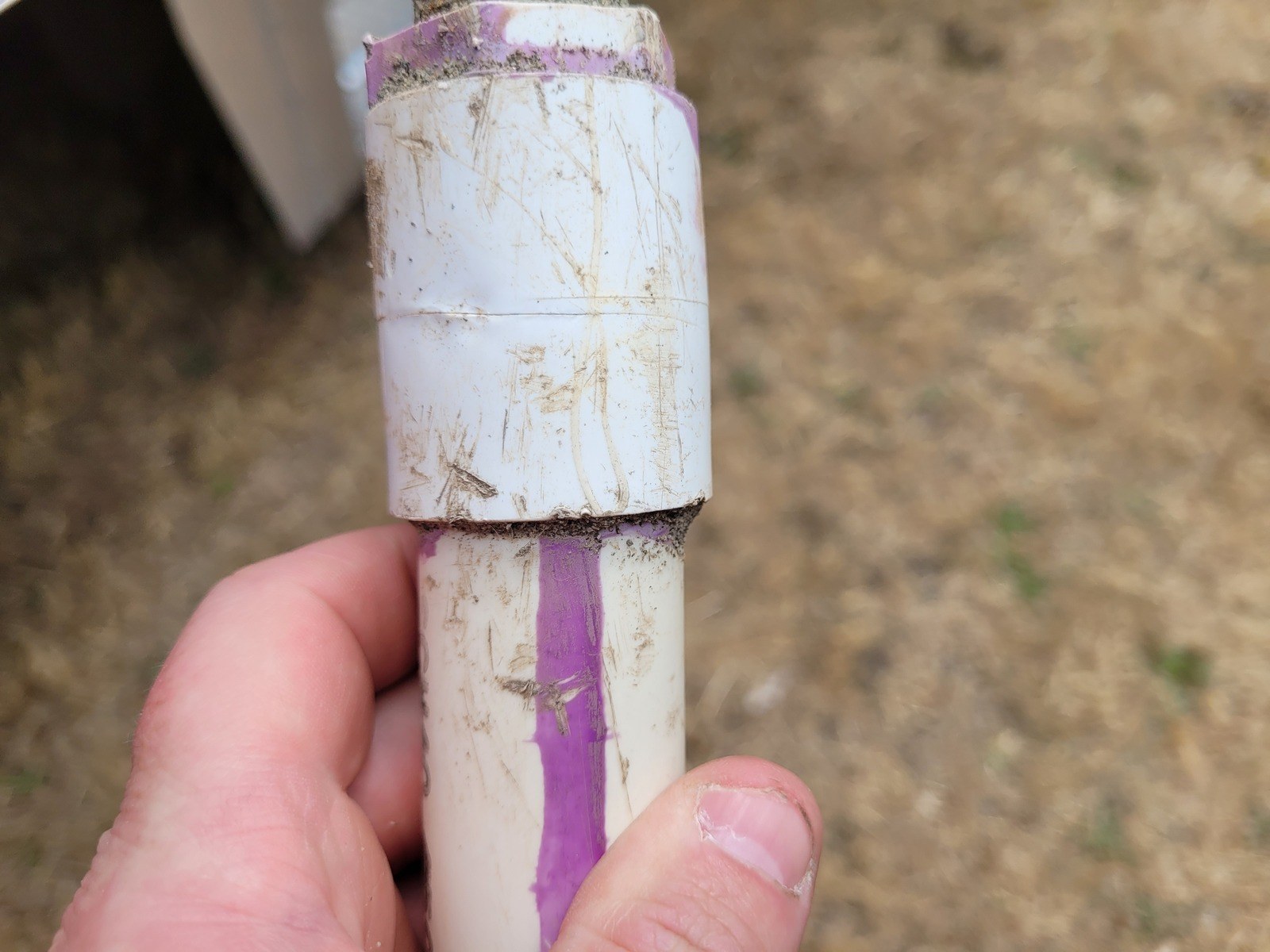 A hand holding a cut-out section of white PVC water pipe with a purple primer stripe, showing a fine crack running around the coupling