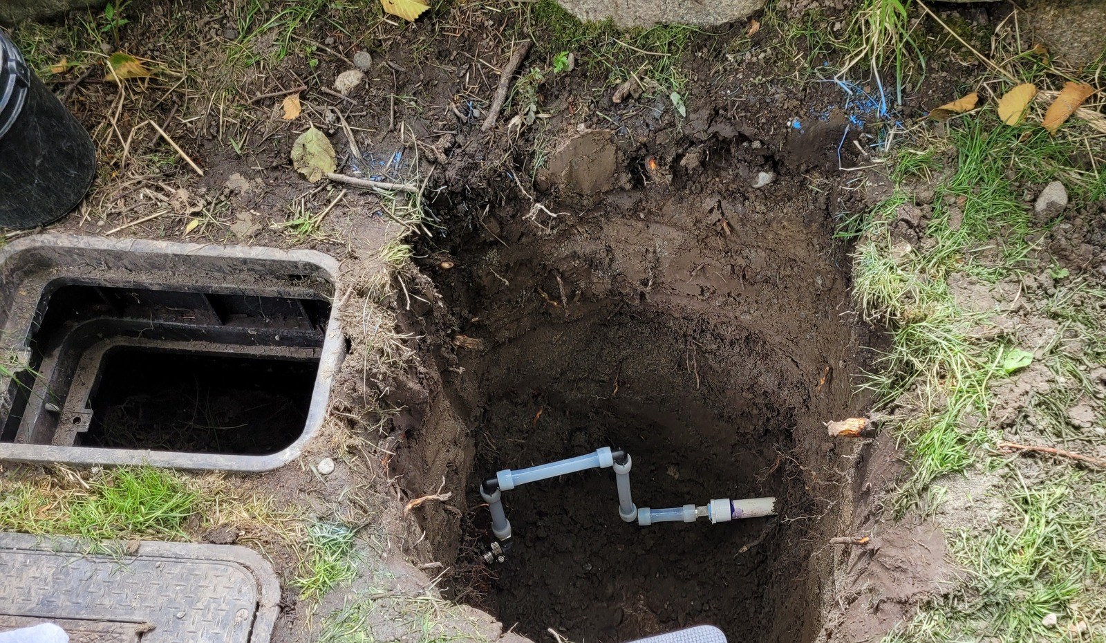 An open hole in a lawn beside a black water meter box, with a new white PVC swing joint of short pipes and elbows in the bottom of the hole